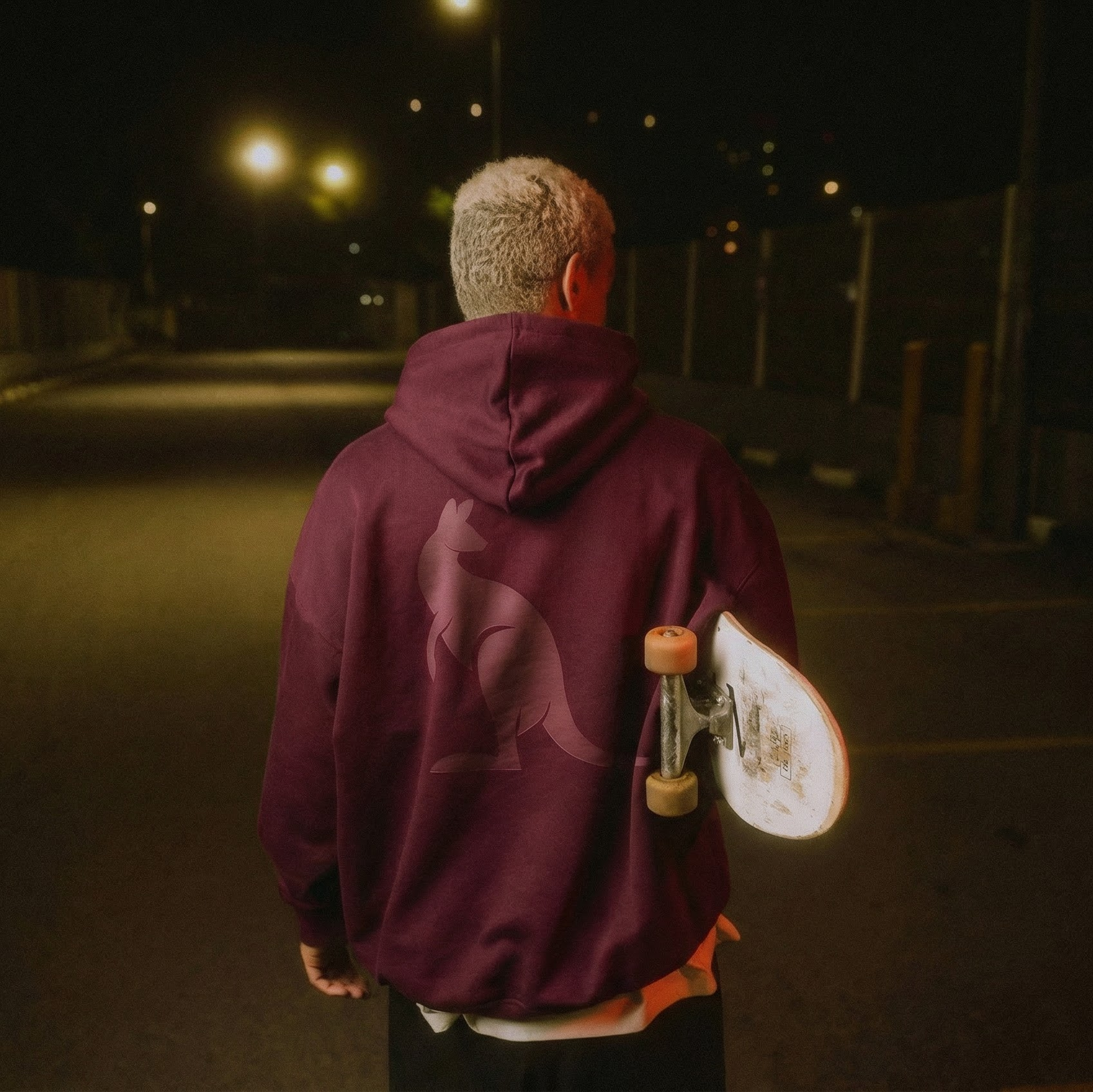 Person wearing a maroon hoodie holding a skateboard on a dark road KANGAR Hoodie System in Oxblood with detachable utility pouch